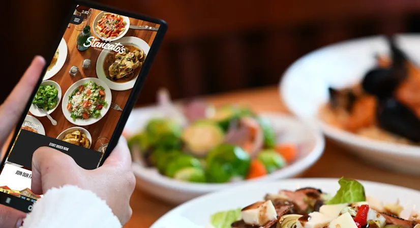 Person sits at a table full of food, with their cell phone out while using the Stancato's app.