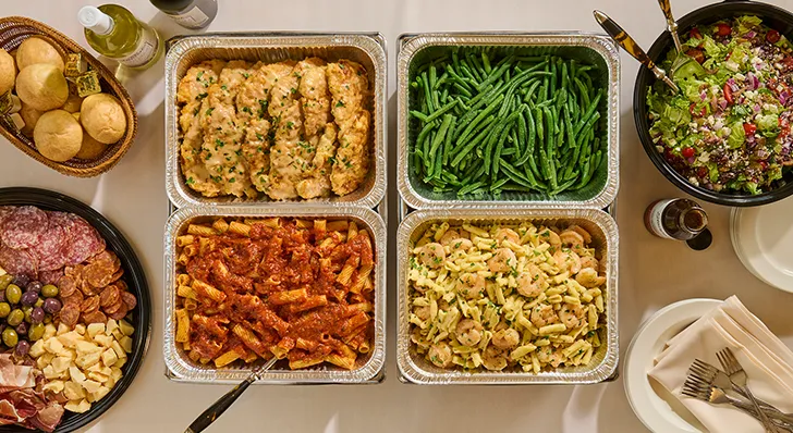 Table full of catered food with chicken, green beans, salad, rolls, charcuterie tray, and shrimp and pasta.