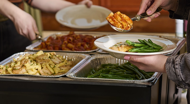 A person serves themself a scoop of rigatoni from a catering table.
