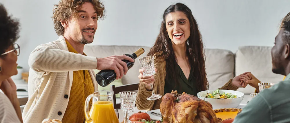 thanksgiving-catering-wine-pairing A cheerful Thanksgiving gathering features friends enjoying a catered meal with wine pairings. A man pours red wine as others smile around a table set with a roasted turkey, salad, and orange juice, creating a warm and inviting scene. The elegant glassware and relaxed atmosphere highlight the perfect blend of gourmet Thanksgiving catering and thoughtful wine pairing.