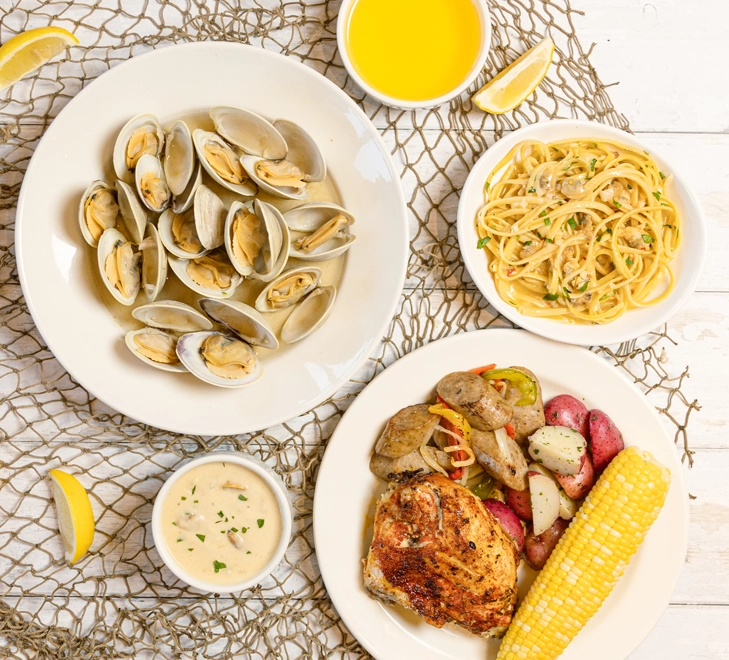 An overhead shot of clams with broth and drawn butter, linguine, chicken, corn and sausage and peppers